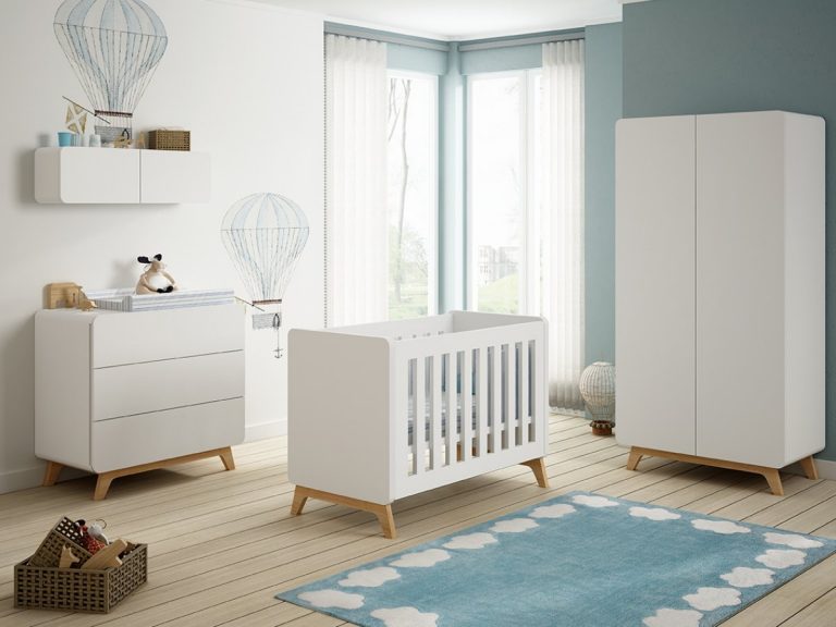 Una habitación infantil bien organizada y bien iluminada con muebles blancos que incluyen una cuna, una cómoda y un armario. Las paredes están decoradas con calcomanías de globos aerostáticos. Una alfombra azul con motivos de nubes blancas añade un toque de color al suelo de madera.